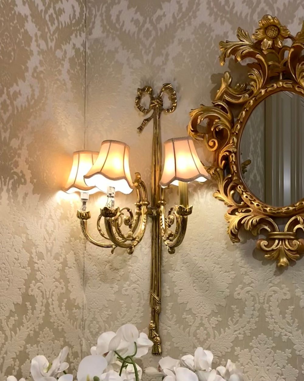 Silver-beige damask wallpaper with a lit brass double sconce, a carved gold mirror and white orchids