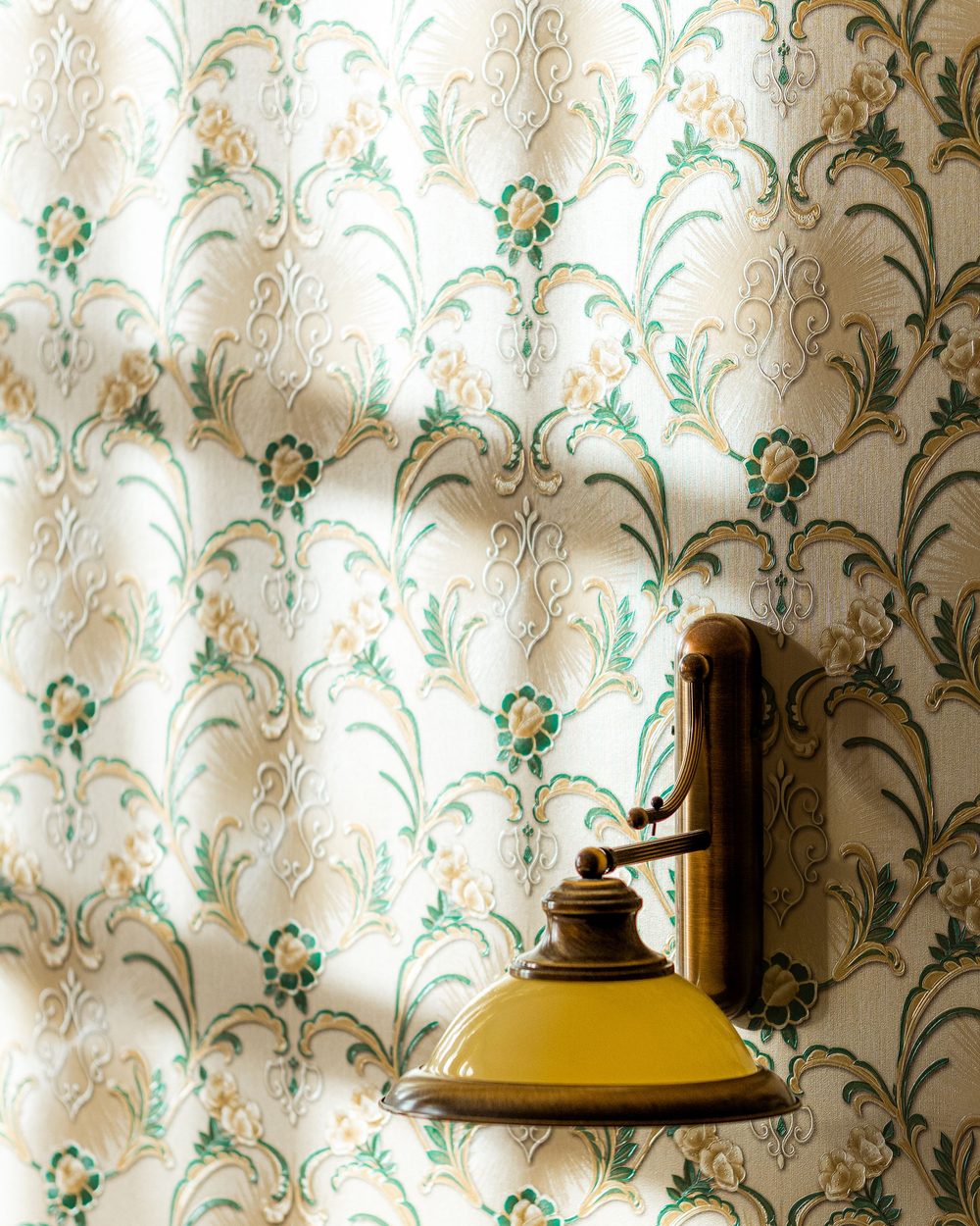 Brass wall sconce on cream and green ornamental wallpaper with soft window shadows