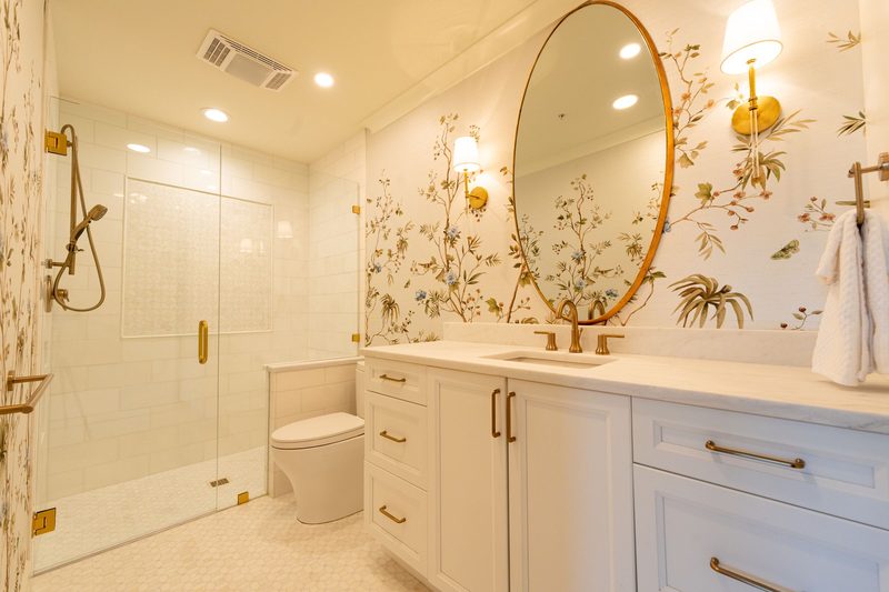 Bathroom with ivory bird-and-branch wallpaper, an oval brass mirror and a glass shower