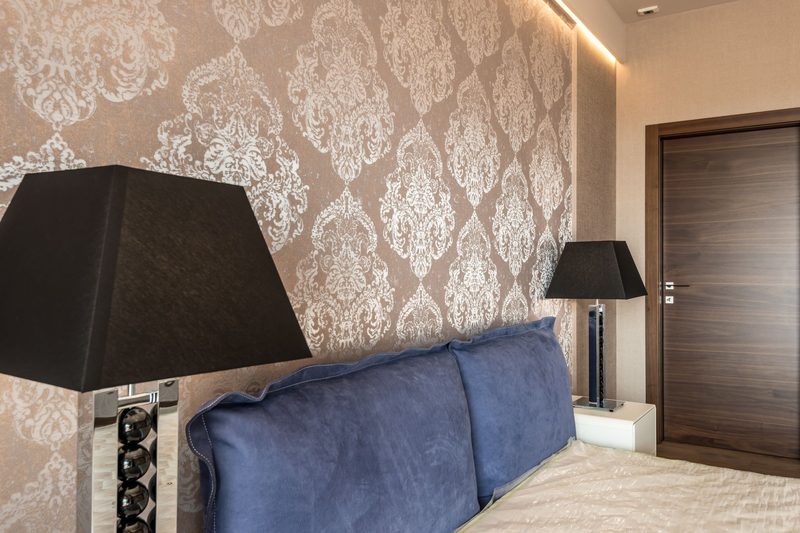 Bedroom with taupe and silver damask wallpaper behind a blue velvet headboard and black lamps
