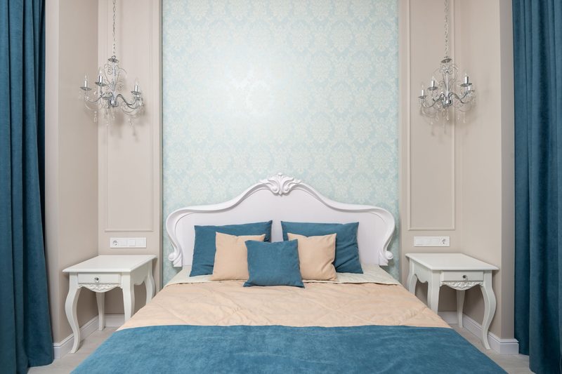 Symmetrical bedroom with a pale damask wallpaper panel behind a carved white bed, crystal sconces and teal curtains