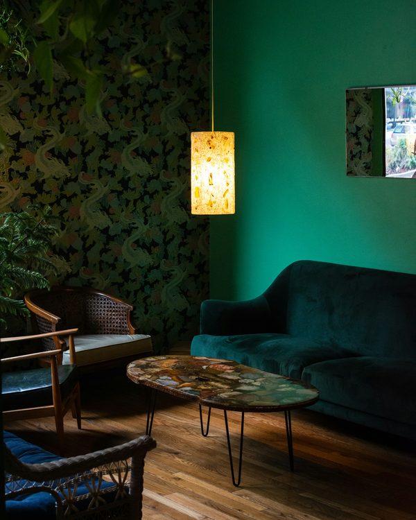 Moody lounge with a dark botanical wallpaper wall beside emerald walls, a velvet sofa and a glowing pendant lamp