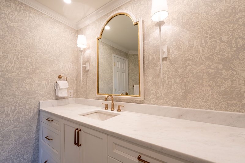 Bathroom vanity with a marble top, an arched gold mirror and grey-beige toile wallpaper