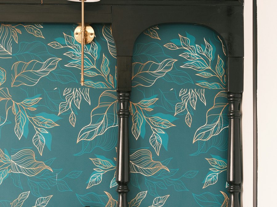 Teal wallpaper with a gold leaf motif framed by dark painted millwork and a brass wall lamp