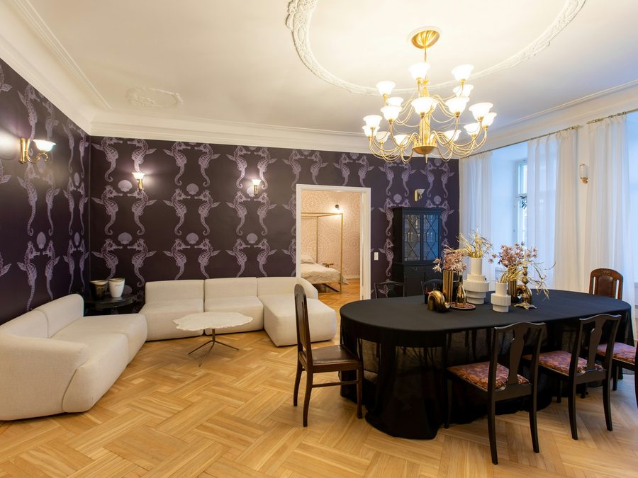 Open living and dining room with deep plum ornamental wallpaper, a white sofa and a brass chandelier